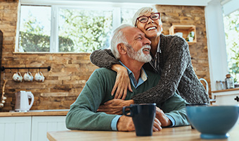 An older married couple hugging in their kitchen.  