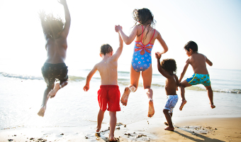 Several children holding hands jumping for joy on a beach.